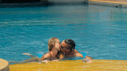 The loving couple hugs and kisses, drinking blue cocktail alcohol liquor in swimming pool at hotel outdoor. Portrait of caucasian man and woman. Creative hairstyles bodybuilder, swimsuit, sunglasses.