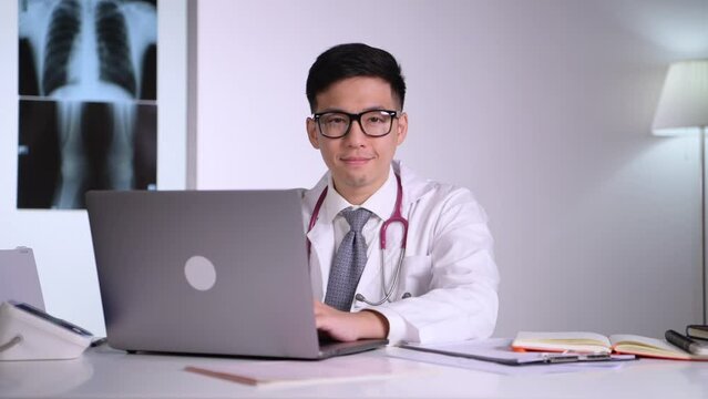 Kind and polite young Asian medical doctor orthopaedic surgeon wearing stethoscope sitting with laptop looking straight with x-ray image behind in examination room in hospital.