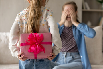 Kid holding, hiding surprise pink gift box behind back, walking to mom. Mother covering eyes with hands, ready to receive birthday present from preschool daughter girl. Close up of wrap, 8 march