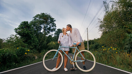 Biking road trip. Love couple on blue bike in white clothes on forest road. Just married woman and man kiss, hugs, stand on bicycle. Wedding, honeymoon. Cycling Cycle Fix. Asia Thailand ride tourism.