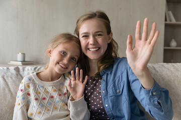 Happy sweet daughter kid and young mom waving hand hello at camera, talking on video call, laughing, smiling, recording video for blog, vlog. Mother and girl home head shot portrait