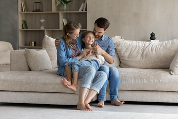 Happy millennial parents holding cheerful daughter kid in arms, enjoying parenthood, leisure time on comfortable couch in living room interior of cozy house. Family relationships, closeness concept