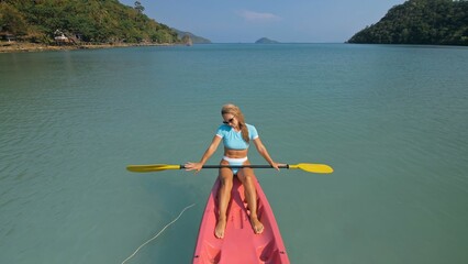 Pretty sportive blonde lady in stylish swimsuit sails on pink plastic canoe along azure ocean bay...