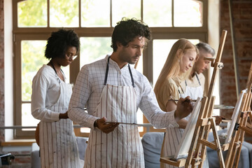 Focused smiling young multiracial people in aprons drawing on canvas with paintbrushes, mixing oil paints on palette, feeling inspired enjoying creative hobby activity at group lesson in loft studio.