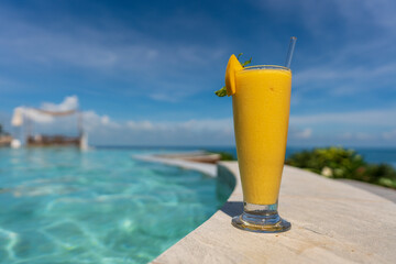 Glass of yellow mango smoothie on swimming pool and sea beach background at summer sunny day. Island Koh Phangan, Thailand