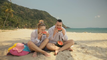 Fototapeta premium The cheerful love couple holding and eating slices of watermelon on tropical sand beach sea. Romantic lovers two people caucasian spend summer weekend in outdoor. Hat, backpack white shirt beachwear.