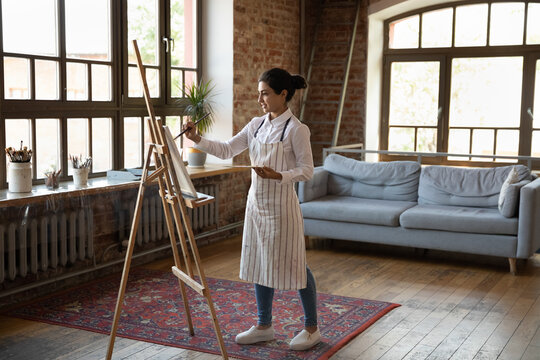 Full length creative motivated millennial Indian female artist in apron using palette with mixed paints and paintbrushes drawing pictures on canvas, standing alone in modern studio or gallery.