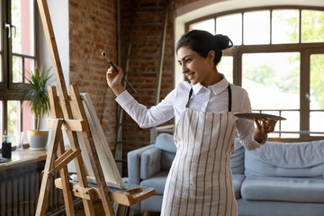 Happy inspired young Indian woman in apron and tied hair holding palette and paintbrush, creating...
