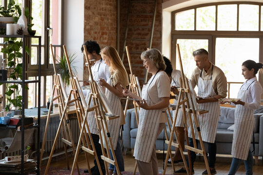 Group of happy multiethnic different generation people wearing aprons holding mixed paints on palette in hands, enjoying drawing pictures on canvas in creative art lesson in modern loft studio.