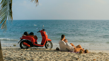 Scooter road trip. Lovely couple on red motorbike in white clothes on sand beach. Just married people kiss hugs walking near the tropical palm trees, sea. Wedding honeymoon by ocean. Motorcycle rent.