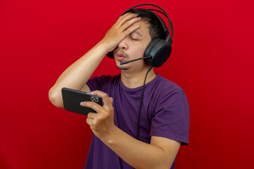 An asian man is playing a losing game on his smartphone and wearing a headset.