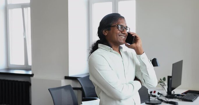 Indian businessman standing at workplace make call, talk to client remotely. Office male employee sales, manager speaks on smart phone solve business working in coworking space. Tech, workflow concept