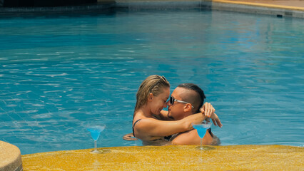 The loving couple hugs and kisses, drinking blue cocktail alcohol liquor in swimming pool at hotel outdoor. Portrait of caucasian man and woman. Creative hairstyles bodybuilder, swimsuit, sunglasses.