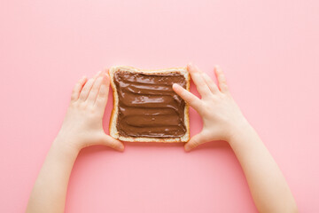 Baby girl hands touching fresh slice of white bread with brown chocolate cream on light pink table background. Pastel color. Closeup. Point of view shot. Sweet snack. Top down view.