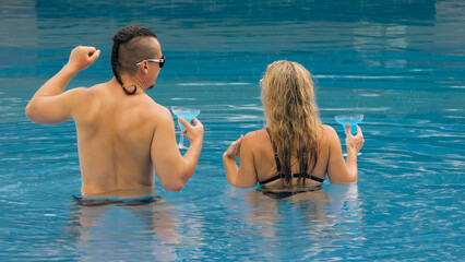 The loving couple hugs and kisses, drinking blue cocktail alcohol liquor in swimming pool at hotel outdoor. Portrait of caucasian man and woman. Creative hairstyles bodybuilder, swimsuit, sunglasses.