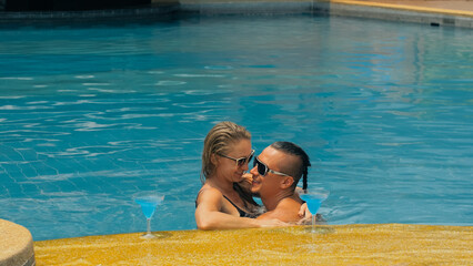 The loving couple hugs and kisses, drinking blue cocktail alcohol liquor in swimming pool at hotel outdoor. Portrait of caucasian man and woman. Creative hairstyles bodybuilder, swimsuit, sunglasses.