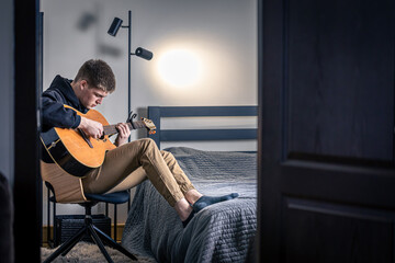 A young man plays the acoustic guitar in his room at home.
