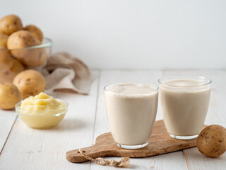Potato milk in glasses on white wooden background. Pouring vegan milk in glass, with potato puree and potato tubers on background. Copy space. Home made potato milk made from boiled potatoes