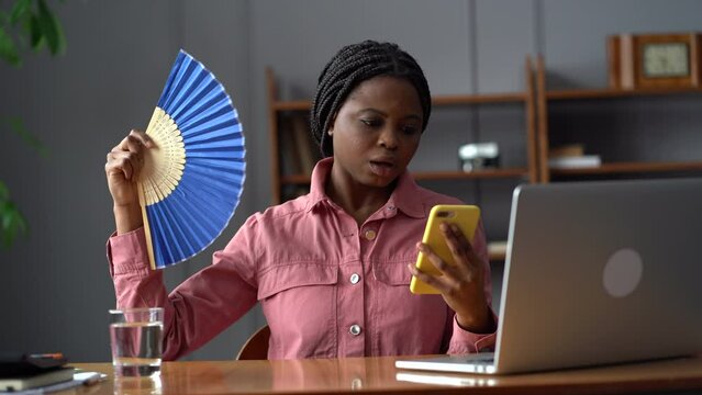Frustrated woman in panic try to contact doctor using mobile phone hold paper fan to relief hot temperature anxiety and tension. Young stressed black female suffer from overheat in office overwhelmed 