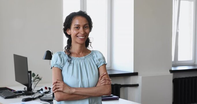 Confident 35s African businesswoman, company staff member, pose alone in office at workplace with arms crossed, smile look at camera. Professional employee, advisory, qualified worker portrait concept