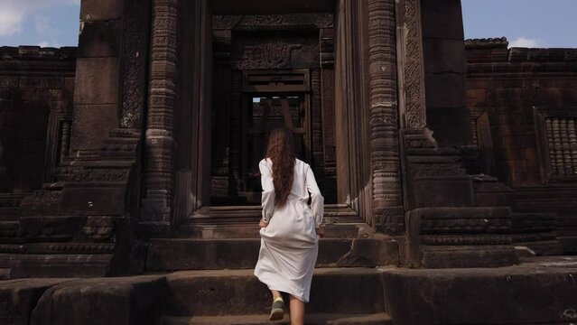 Young beautiful woman in long white dress going up the stairs to palace in Wat Phou – ruined Khmer Hindu Temple complex. Champassak, Laos, Asia. Ancient culture religious architecture, ruin. Slow