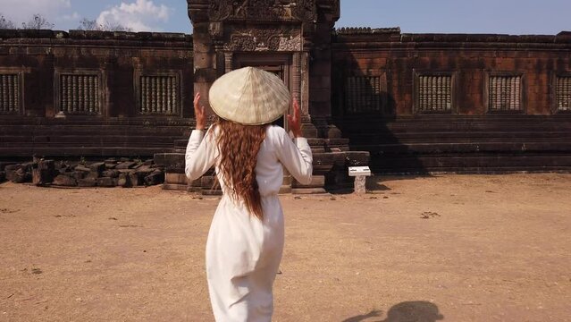 Young beautiful woman in long white dress and vietnamese hat going to palace in Wat Phou ruined Khmer Hindu Temple complex. Champassak, Laos, Asia. Sunny. Ancient culture religious architecture. Slow