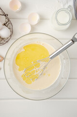 Cooking dough for traditional thin pancakes or crepes. Eggs, flour, salt, sugar, milk and butter in a bowl on a white wooden background. Recipe step by step. Food for Maslenitsa.