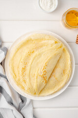 Stack of traditional thin pancakes or crepes on a plate with honey and sour cream on a white wooden background. Food for Maslenitsa. Copy space.
