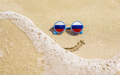 Fototapeta na wymiar Sunglasses with flag of Russia on a sandy beach. Nearby is a sea lightning and a painted smile. The concept of a successful vacation in the resorts of Russia..
