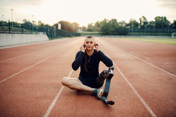 Fototapeta premium A runner with artificial leg sitting at the stadium, using headphones and relaxing.