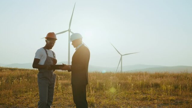 Two partners standing on field with wind turbines. Engineer and inspector working together on windmill farm. African american engineer and indian inspector using digital tablet during meeting outdoors
