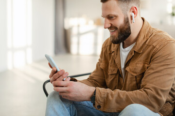 Side View Of Cheerful Guy Using Smartphone Wearing Earbuds Indoor