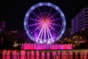 Long exposure photo of a Ferris wheel ride in operation behind water fountain jets in the city, surrounded by palm trees and beautifully colored buildings on fair night. Fun. Entertainment.
