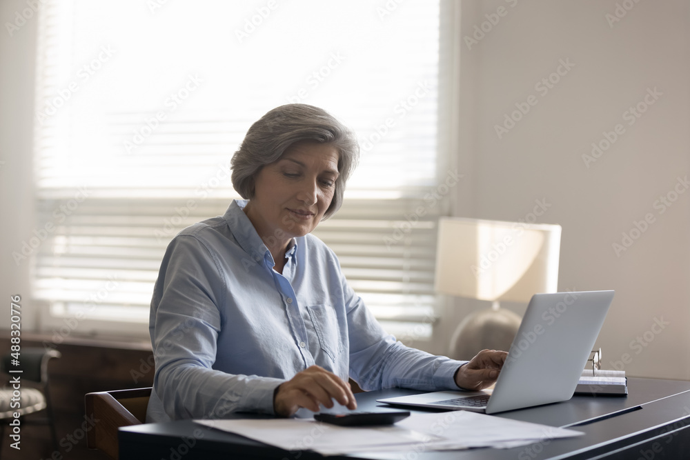 Wall mural Pleasant happy middle aged senior woman calculating utility bills or taxes, involved in managing incomes or outcomes using laptop apps, paying for services, planning expenditures, accounting.