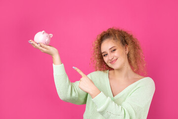 A cute young girl with curly red hair holds a piggy bank, a pink piglet in her hands. The concept of wealth and safety of money.