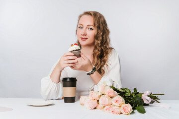 A beautiful young woman with curls on a white background holds a cupcake with cream and berries and a plastic cup of coffee or tea in her hand.