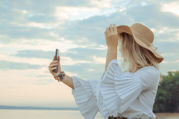 Portrait of a girl taking a selfie on the smartphone. Young beautiful 30 years woman caucasian appearance in straw hat with satin ribbon bow. Copy space. Summer lifestyle concept. Natural background