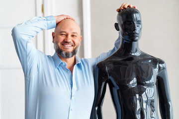 Bald no hair glad adult man with beard smiling teeth, holding head and top of naked artificial plastic black manikin. Light indoor cabinet office. Hair transplant replacement treatment service. Close