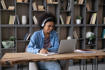 Happy beautiful millennial African American woman in headphones looking at laptop screen, watching lecture or webinar online, holding video call with mentor, preparing for exams, writing notes.