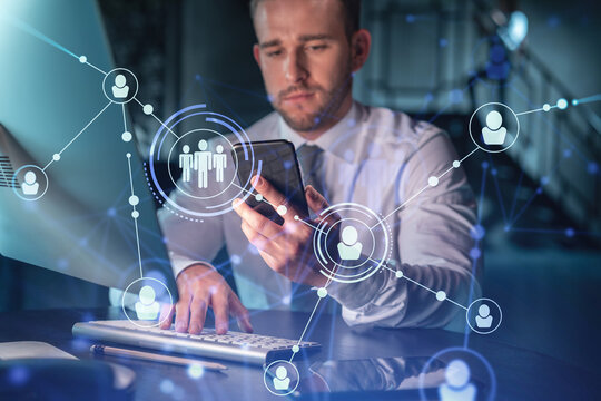 Businessman in casual wear using smart phone to check new candidates for international business consulting. HR, social media icons over modern office background at night time.