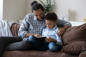 Happy bonding affectionate young African American father and small child boy using digital computer tablet, playing online video games, watching funny cartoons resting on sofa, tech addiction concept.