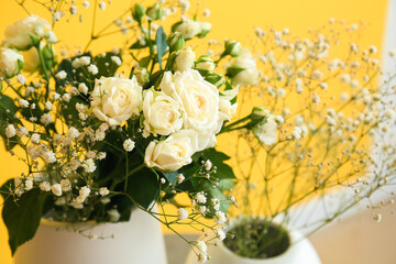 Vases with roses and gypsophila flowers near yellow wall, closeup