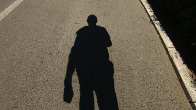 A shadow on the asphalt road of a person roller skating and filming himself with a sports action camera. High angle point of view shot
