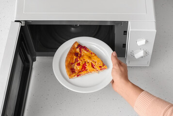 Woman putting plate with piece of pizza into microwave oven on table, closeup - obrazy, fototapety, plakaty