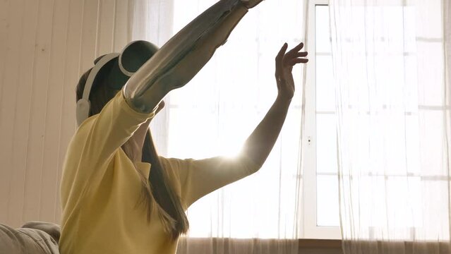 Woman with prosthetic arm enjoys virtual reality technology in sunny room. Lady in goggles plays game gesticulating. Lifestyle of disabled people