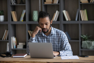 Pensive young African American businessman looking at laptop screen, stuck with difficult task, considering problem solution, preparing presentation. analyzing sales statistics data, working at office