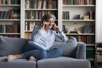 Full length happy beautiful middle aged old retired woman holding phone call conversation with friends or family, lying on comfortable couch, discussing life news, older people and modern technology.