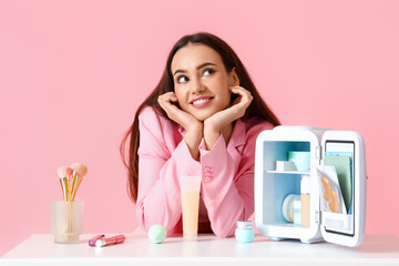 Beautiful young woman with small refrigerator and cosmetic products sitting at table on color background