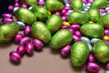 Small pink, yellow, green and silver foil wrapped chocolate easter eggs with larger lime green eggs, against a pale wood background.