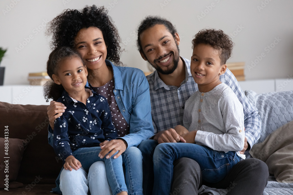 Wall mural Portrait of smiling affectionate young African American couple parents holding on laps cute laughing little children siblings, happy friendly full family looking at camera, relations concept.